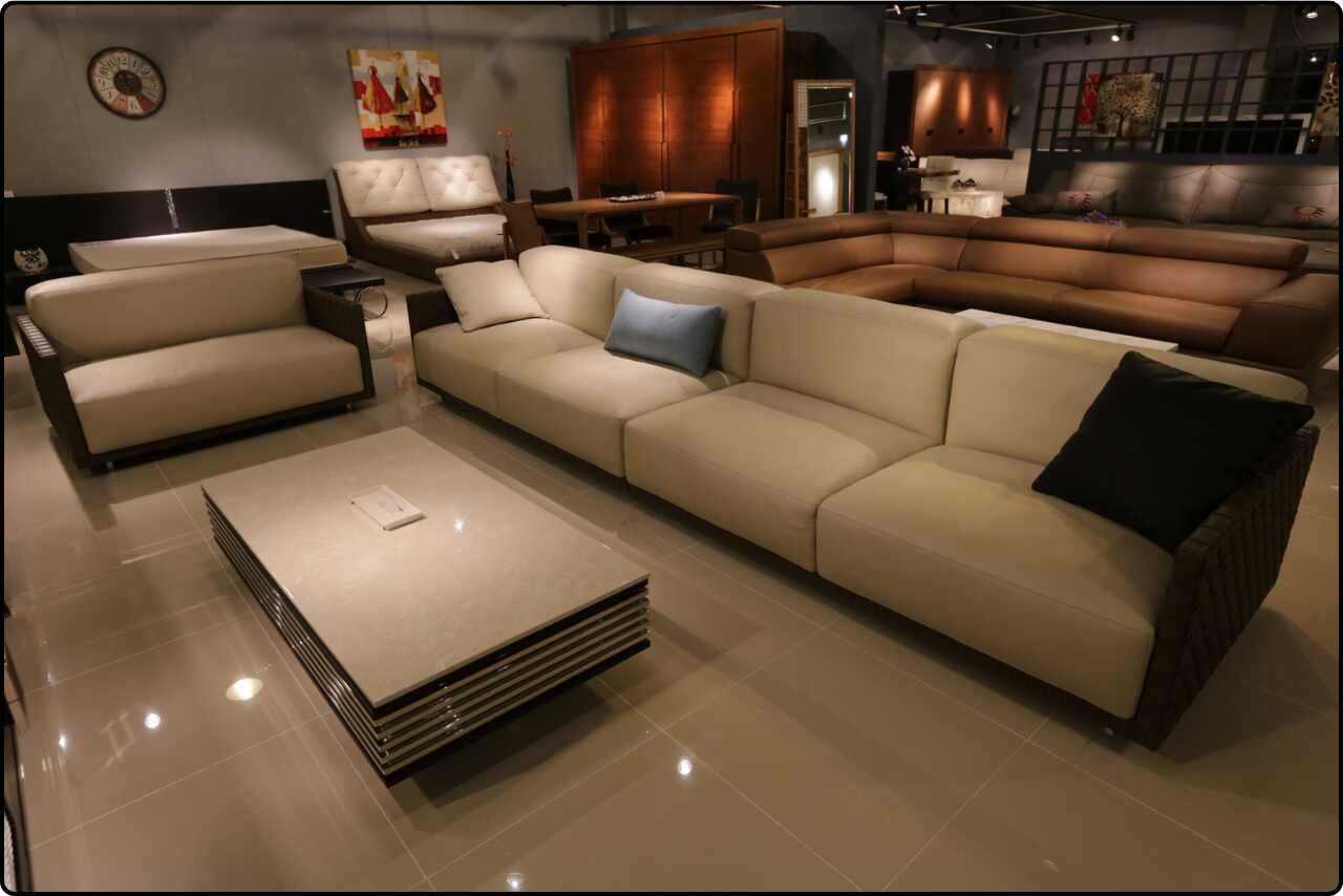 Modern sofa set on display inside a furniture store, featuring a minimalist white sofa with gray and blue accent pillows and a simple wooden coffee table.